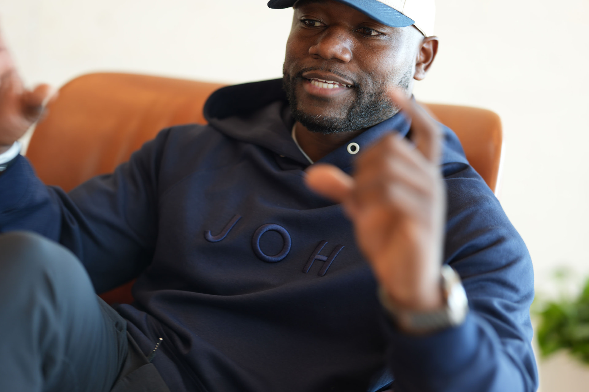 A man in a navy JOH hoodie and cap gesturing while seated in an orange chair